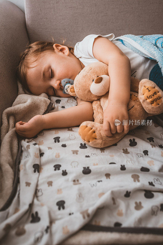 可爱的健康的蹒跚学步的小男孩在床上的毯子下睡觉/打盹，同时拥抱着泰迪熊